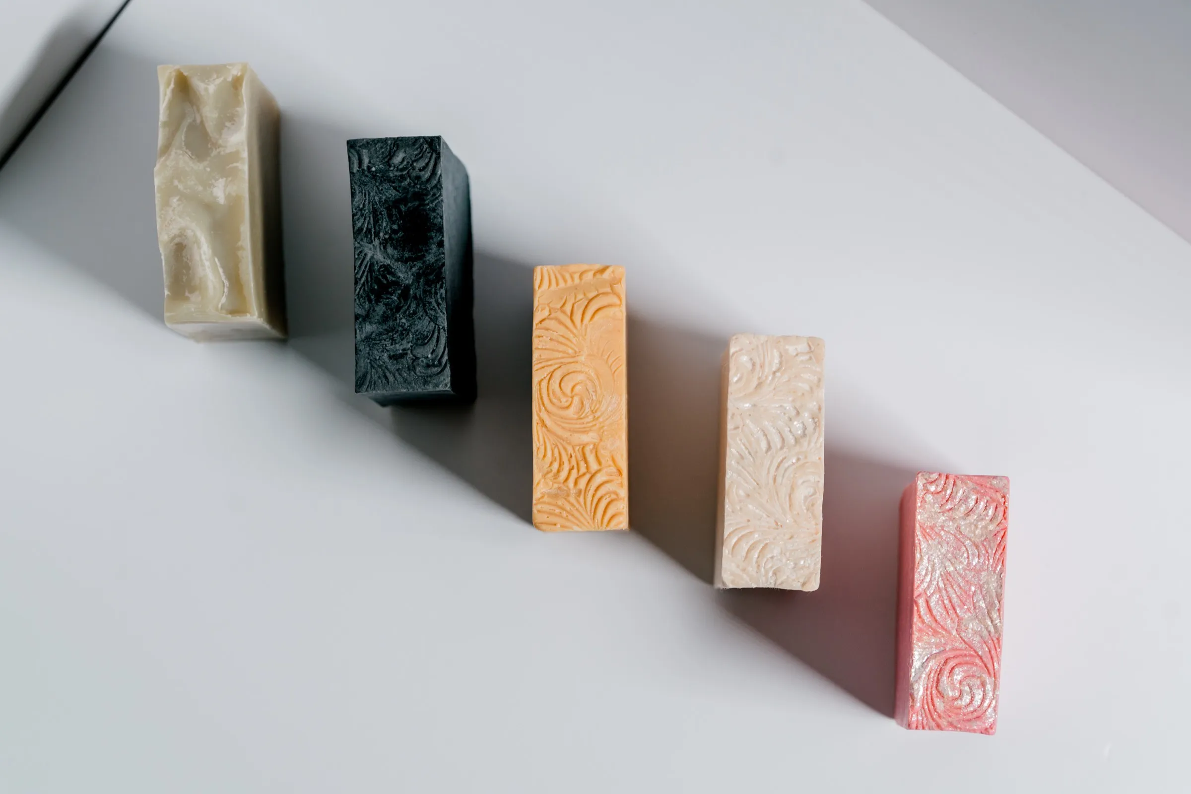 Five handmade bars, olive, charcoal, apricot, ivory and coral, standing in a diagonal row on a white studio table