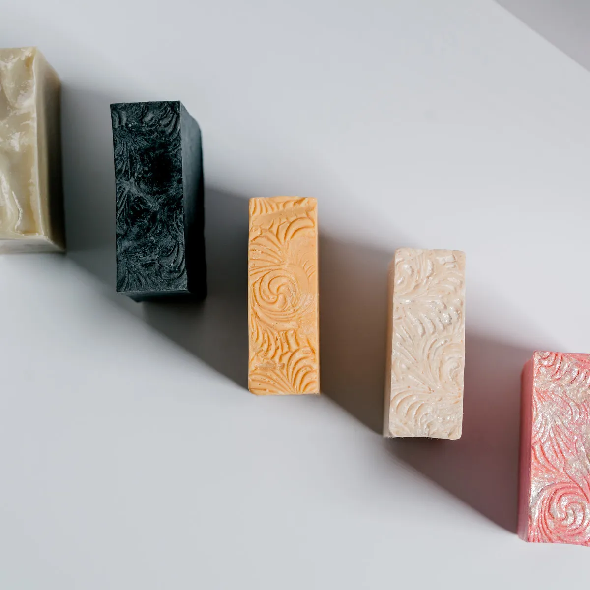 Five handmade bars, olive, charcoal, apricot, ivory and coral, standing on a grey studio ground